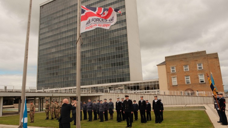 This image shows the Armed Forces Day flag being raised in front of military personnel onlookers