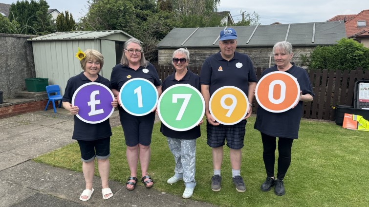 In this photo five volunteers from the group are standing in a line. Each is holding a large circular board  with one of each of the figures £1790 on them.