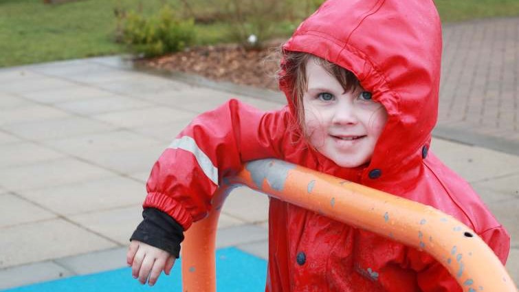 this is a photo of a nursery pupil wearing a red hooded jacket. She is pictured in the playground of the school.