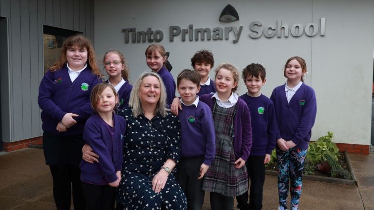 Tinto Head Teacher Alex Stark is pictured at the front door of the school surrounded by eight of the school's pupils. The school name can be seen in the background.